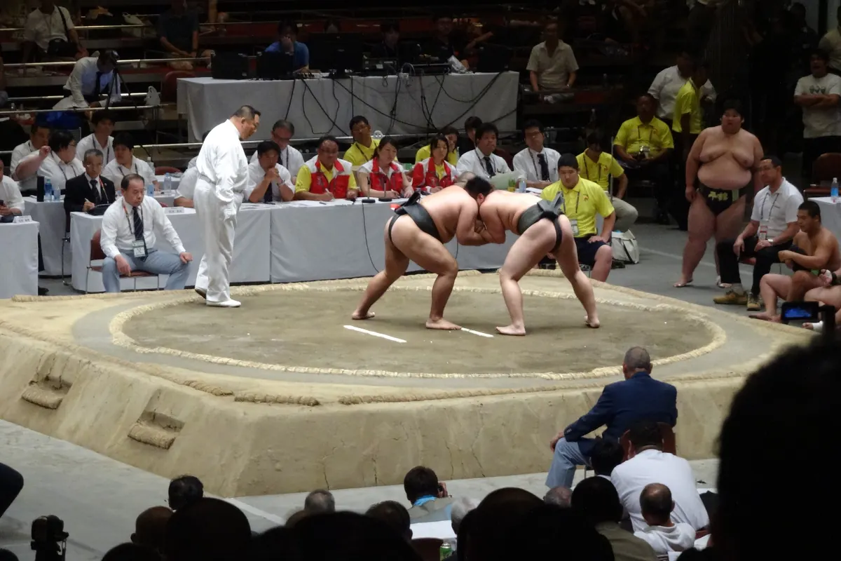 Dva amatérští sumo zápasníci se chytí v hliněném ringu, zatímco rozhodčí v bílém dřepí vedle nich a lidé sedí za nimi u dlouhého stolu