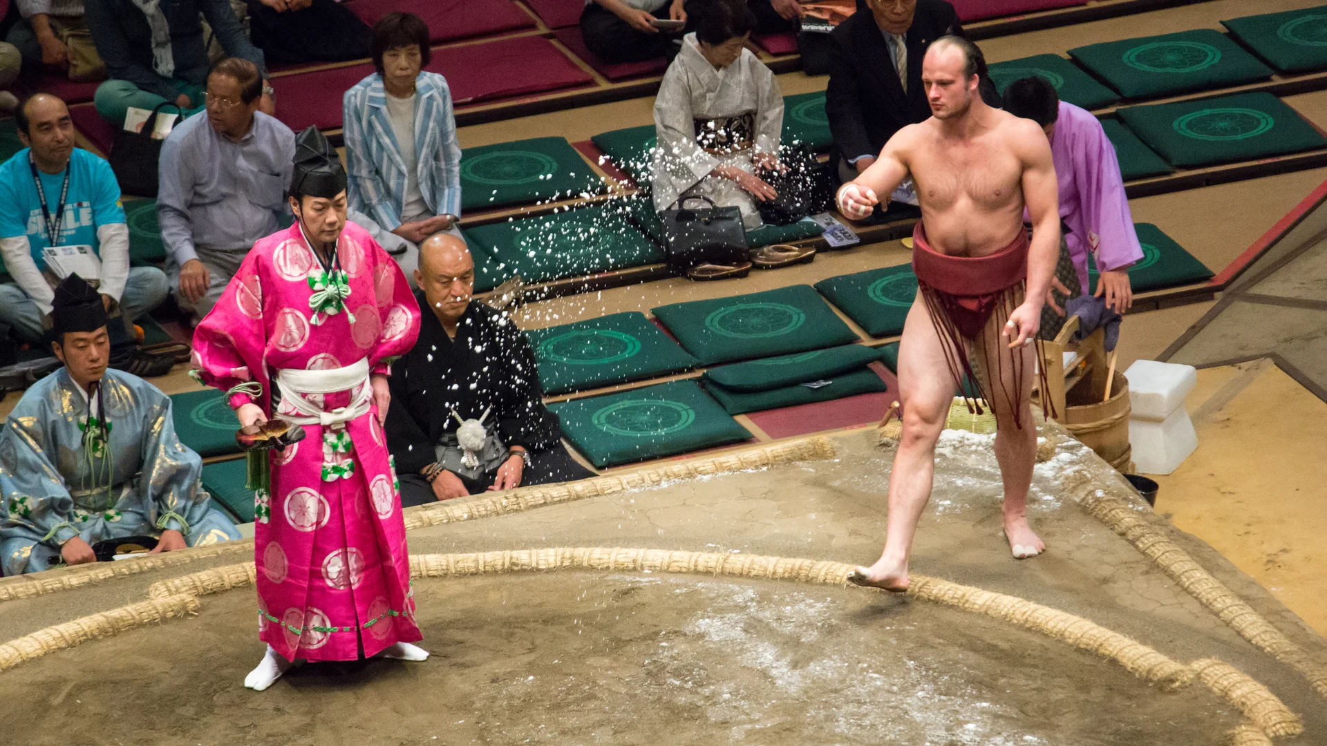 En sumobryder i rødt bælte kaster salt ind i en lerring ved en turnering i Tokyo, mens en dommer i lyserøde klæder står ved siden af ham